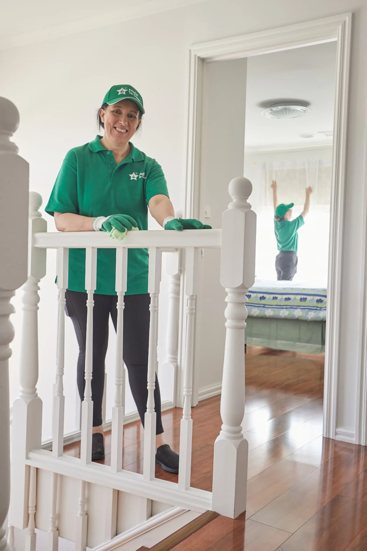 Smiling Maid to Clean cleaner polishing a stair banister while a colleague cleans a bedroom window